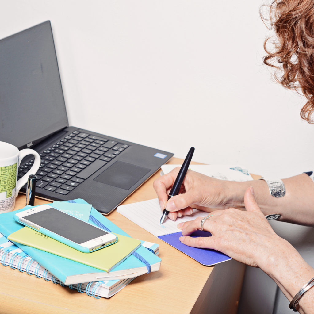 A lady writing using a fountain pen in front of a laptop. with notebooks at the side.